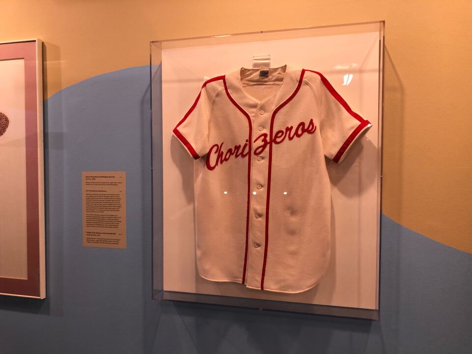 A baseball jersey made of white material with red striping is in a clear case on the wall. The words Chorizeros are written in red stitching across the chest. 