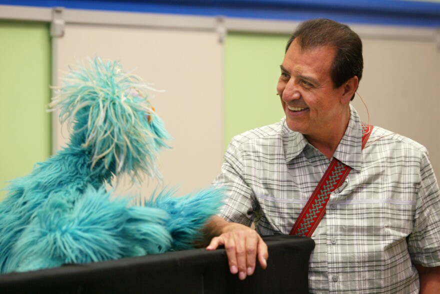 Emilio Delgado wears a plaid, short sleeve collared shirt as he chats with a blue puppet to his right.