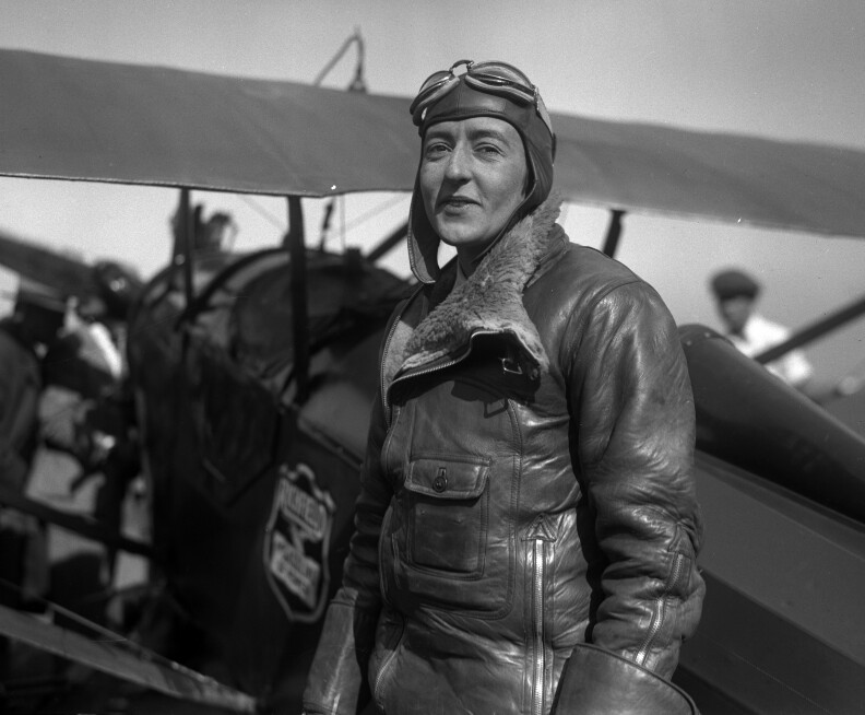 A black and white photo of a fair-skinned woman dressed in a leather aviator jacket, standing in front of a small airplane. She is facing the camera with a bemused look.