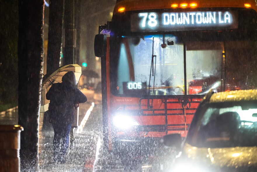 On the left of frame a person with a black coat and tan umbrella walks towards an orange bus that reads "78 Downtown LA." The light from the bus shines on the heavy rain in the air.