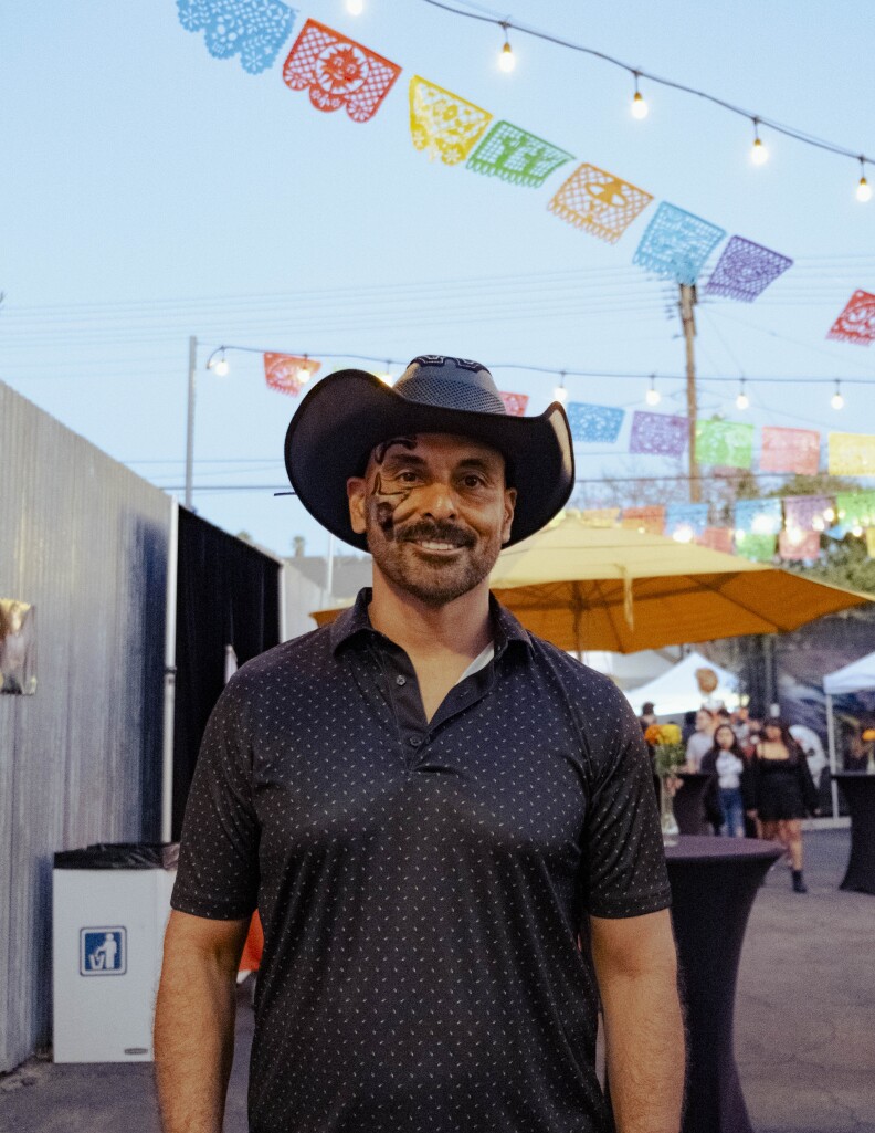 A man with a medium skin tone poses for a photo. He's wearing a black dotted polo and black cowboy hat. He has skull facepaint on and facial hair. In the background are string lights and papel picado.