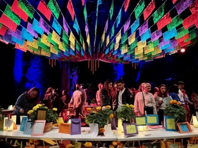 Rows of papel picado, colorful paper flags, hover at night above a massive three-tiered altar with people gathered around, some signing notes to loved ones to honor Dia de los Muertos. 