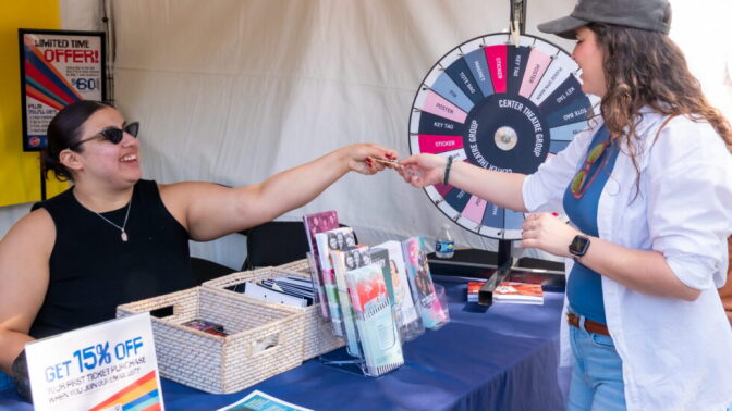 A person hands a raffle ticket to another person standing beside a prize wheel labeled 'Center Theatre Group' at an outdoor booth.