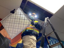 A man in a bright neon yellow safety vest stands on top of a ladder near the ceiling of a hallway while holding a large air filter, perhaps 2 feet long by 2-and-a-half feet wide.