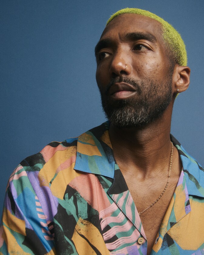 a medium brown skinned man with neon green hair poses in a picture with a blue background. 