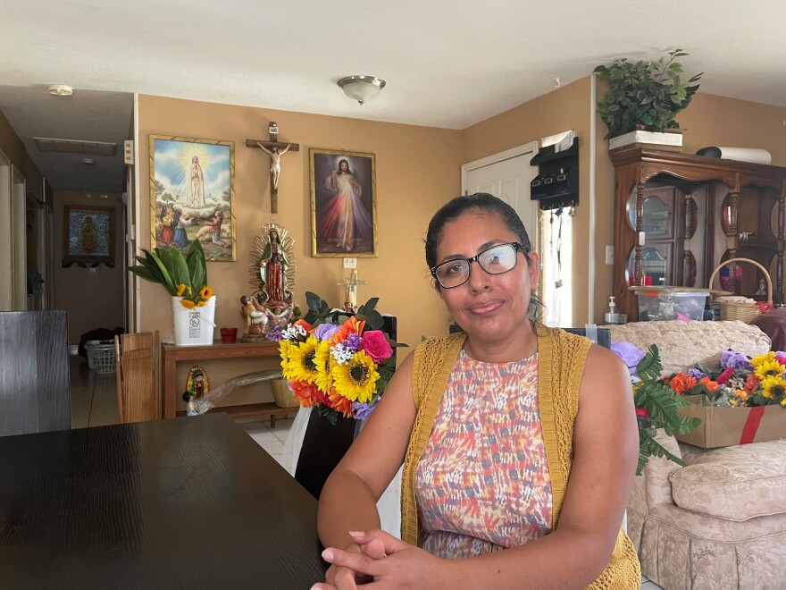 A woman wearing glasses and a knitted vest, sits with her hands folded on a dining table.