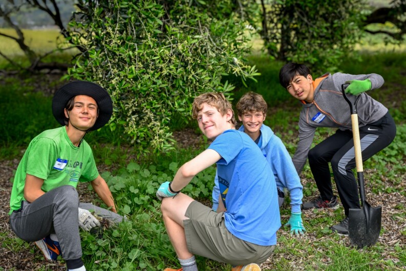 Four teen boys pause to smile for a photo in the middle of volunteering to clean-up an outdoor park. They're all working in the grass and shrubbery. One young man has a shovel and another is wearing a black sunhat.