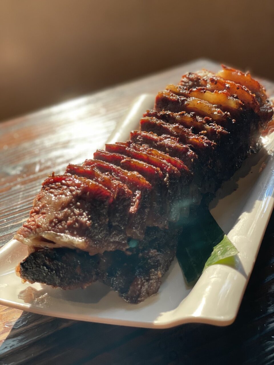 A long line of shortrib beef slices glistens with fat. It sits on a white plate on a brown wooden table.