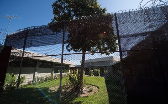 Looking through a mesh metal fence with razor wire on top. On the other side, people in neon green jail clothes are walking and standing outside of a building. 