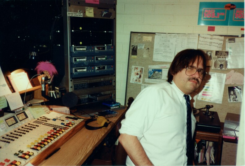 MARS FM Radio DJ Freddy Snakeskin, inside the broadcasting booth at the MARS FM station. On the top of the console there is a troll doll with pink hair. Freddy has longish hair and glasses along with a mustache. He has a sly look on his face like he's questioning the intention of the photographer.