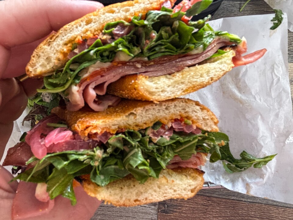 A deli sandwich cut in half, showing layers of sliced cured meat, pickles, crushed potato chips, melted cheese, and a reddish sauce between toasted sandwich bread.