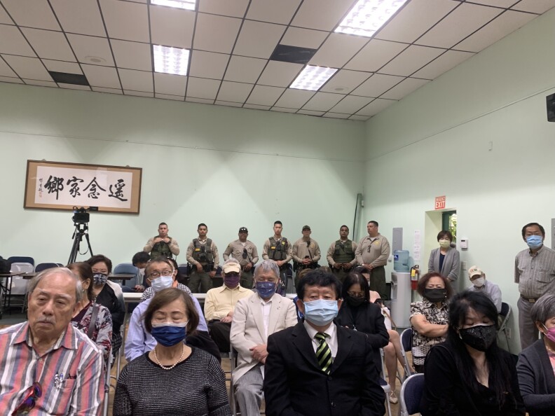 A roomful of mostly older Taiwanese American men and women sitting on folding chairs listening to a news conference. A row of seven sheriff's deputies stand in the back.