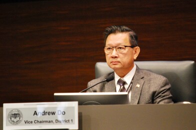 A man in a chair wearing a suit jacket, tie and glasses looks forward with a microphone in front of him. A sign in front has the official seal of the County of Orange and states "Andrew Do, Vice Chairman, District 1."