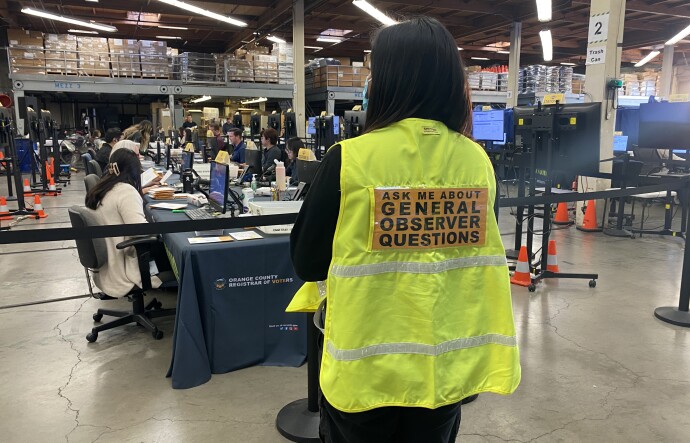 In the foreground, a woman with her back turned wears a bright yellow vest with a sign that reads "Ask me about general observer questions." In the background, a row of people sit in front of computers. 