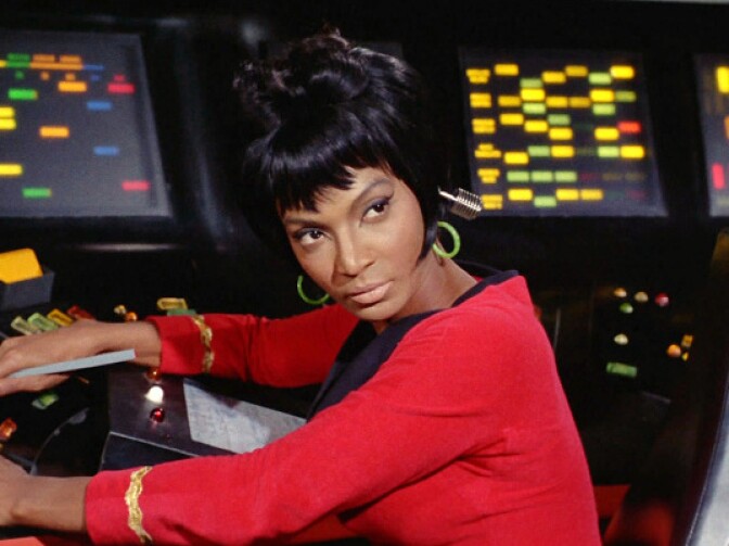 Nichelle Nichols wears a red uniform in front of a control panel on the Enterprise