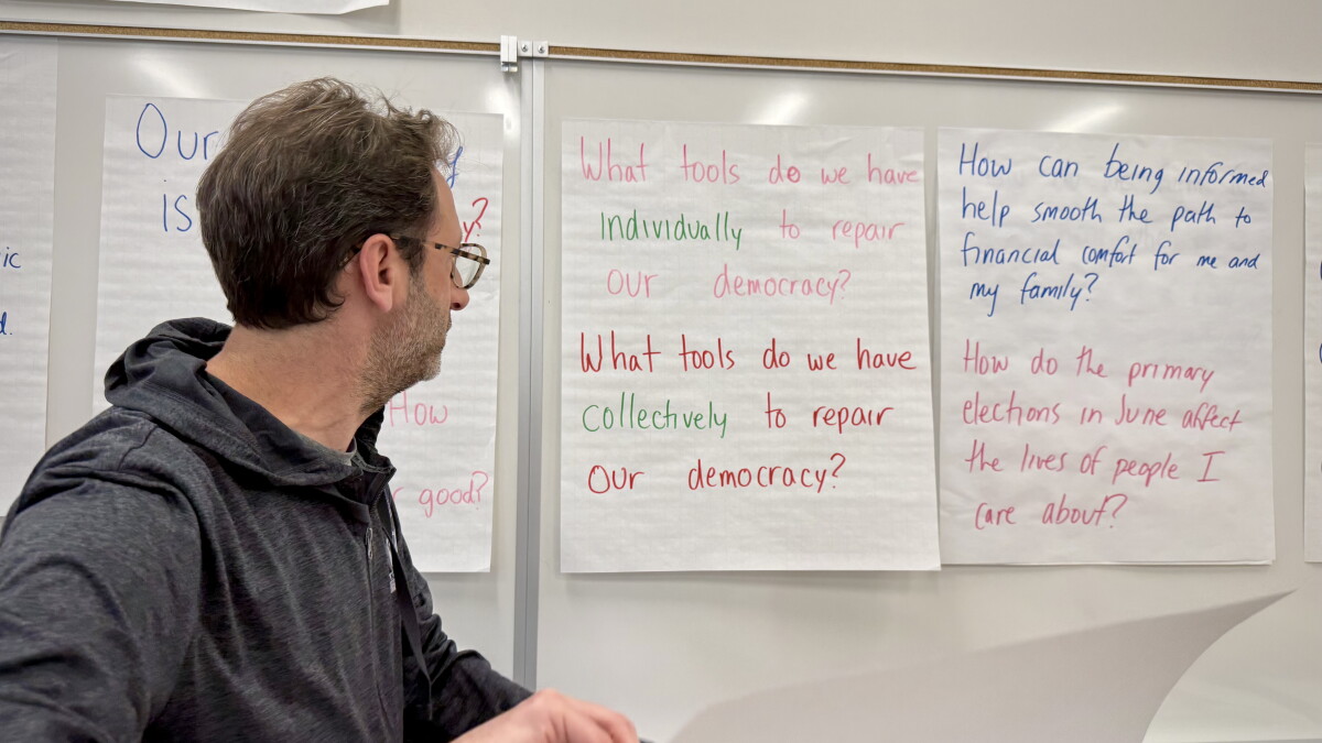 A man with light skin tone and short brown hair wearing a gray hoodie stands in front of several large pieces of paper tacked to a white board. The center sheet reads "What tools d