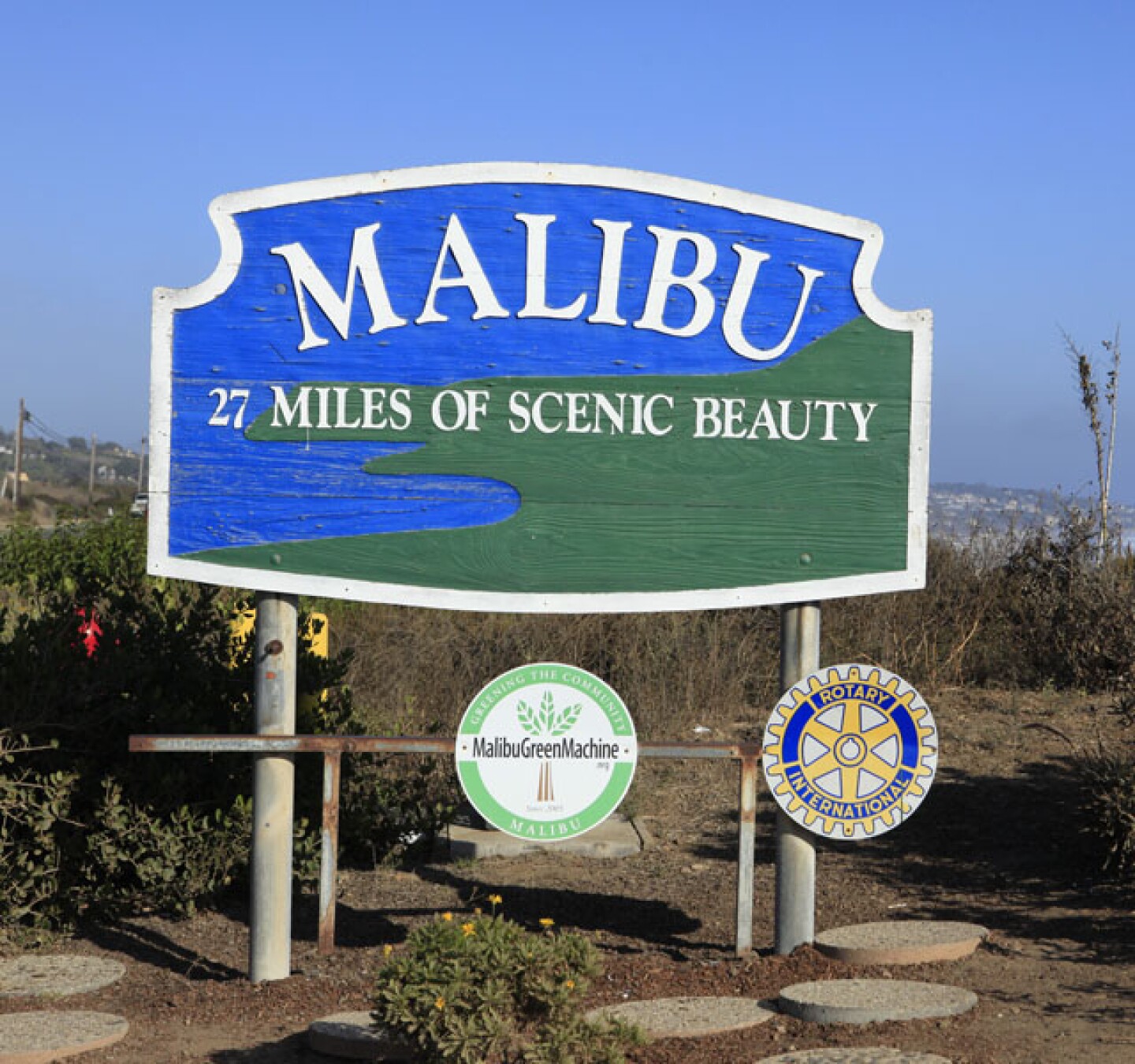 A roadside sign reads Malibu, 27 miles of scenic beauty, emblazoned on a blue and green background depicting the ocean and the shore. Two circular signs are posted below the main sign, one reading Malibu Green Machine and the other a yellow and blue sign for Rotary International.