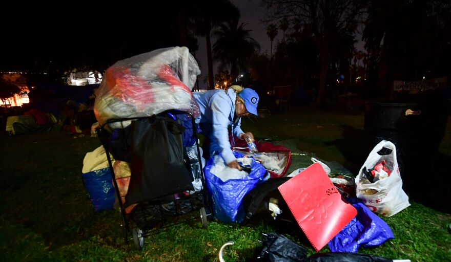 A homeless woman in a blue hat bends over to gather together piles of her belongings, including a large suitcase and a plastic bag.
