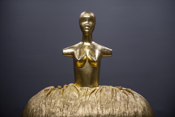 Image is a Gold torso and head on top of a gold skirt. The torso features no arms, breast and no eyes but lips, and nose for face details. Behind a black background.