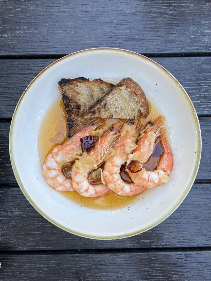 A plate featuring four whole shrimp sit atop two pieces of grilled bread, surrounded by a pale broth. 