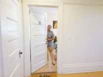 Tisha stands inside her apt while holding the front door open. She is wearing a gray t-shirt and blue jean shorts and sandals.