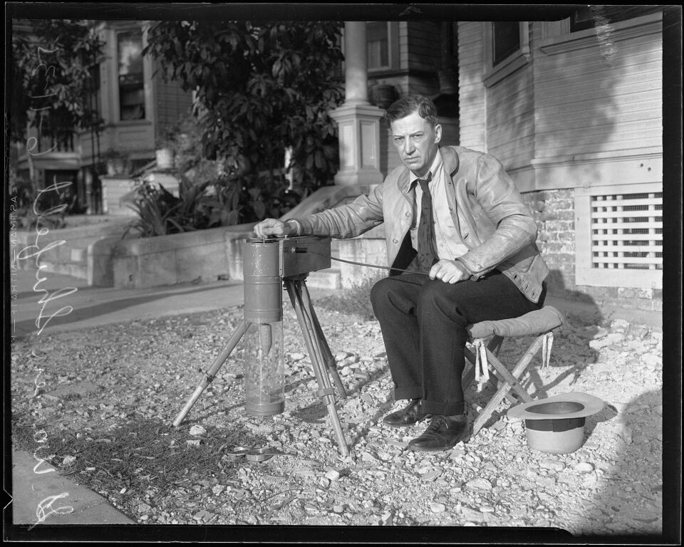 George Warren Shufelt, a white man in a leather jacket and tie, seated on folding stool near a building. His hat is near his feet, while he poses with a cylindrical, tripod-mounted radio X-ray device.