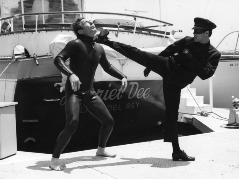 A still from a black-and-white TV show with a masked man in a chauffeur's uniform kicking another man in the chin.