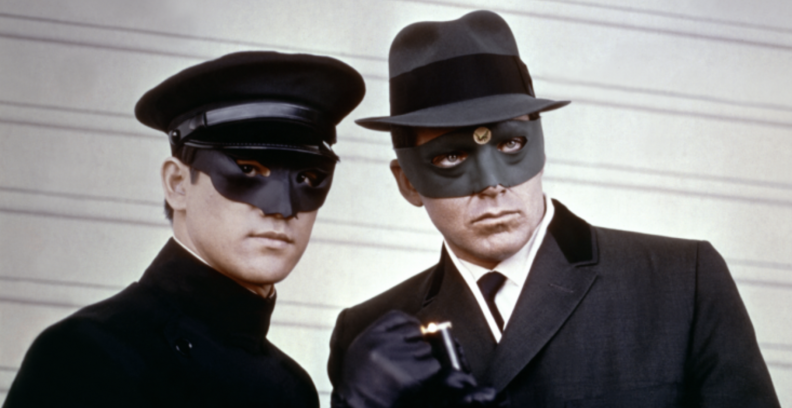 A still of two masked men, one dressed as a chauffeur, the other in 50's-style businesswear.