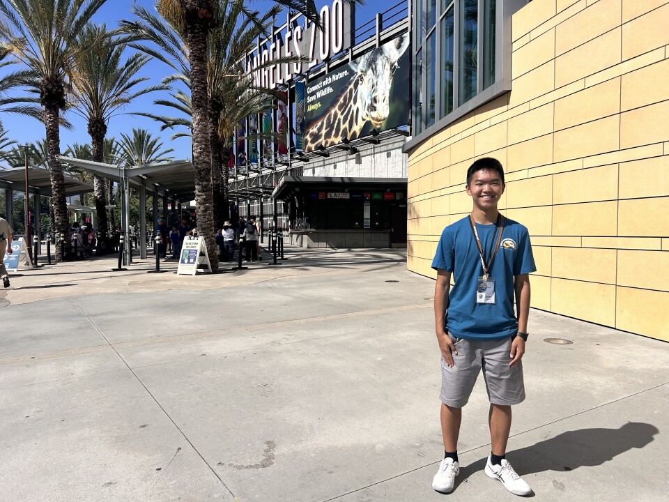 A teenage boy in a blue t-shirt and gray shorts is smiling in front of the exterior of the Los Angeles Zoo. Both hands are to his side and he's wearing a lanyard with a pair of name tags pinned to it.