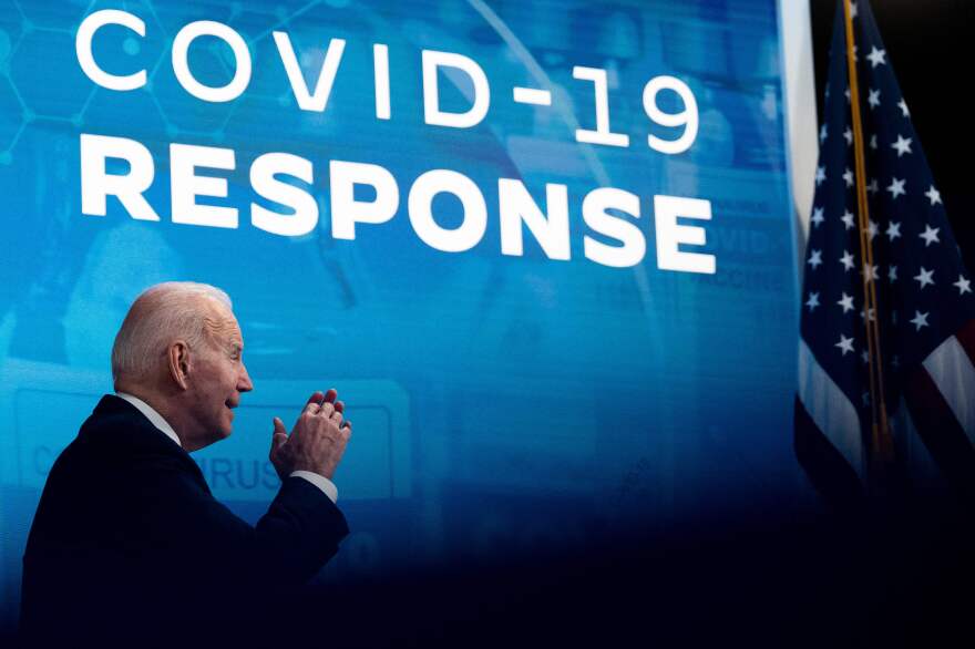 A side view of President Biden with his hands raised in mid-speech. By him is a United States flag and the words "COVID-19 Response" on a blue backdrop.