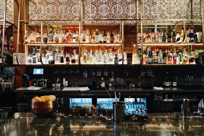 A back-lit bar contains various bottles of alcohol, with rows of cocktail glasses underneath. In front, a large metal-topped bar sits.  