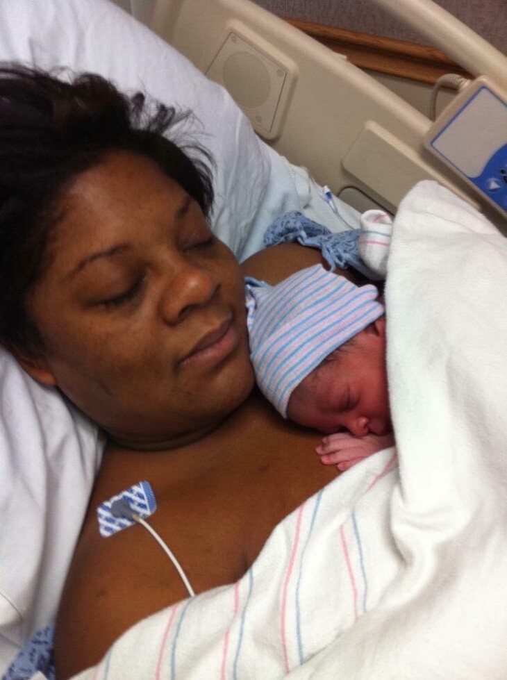 A Black woman rests on a hospital bed, her eyes closed. An infant rests on her shoulder.