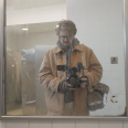 A male presenting man with glasses in a tan jacket looks at himself in a bathroom mirror with a camera pointed towards the mirror.