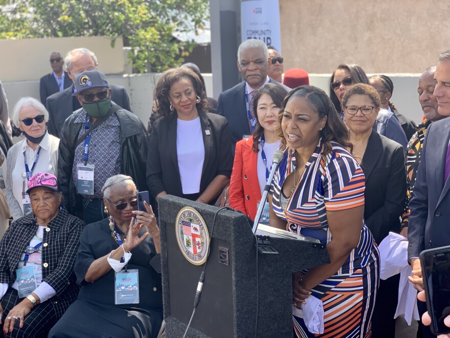 A crowd of more than a dozen people surround an outdoor podium where a woman wearing a multicolored dress stands a microphone during a press conference.