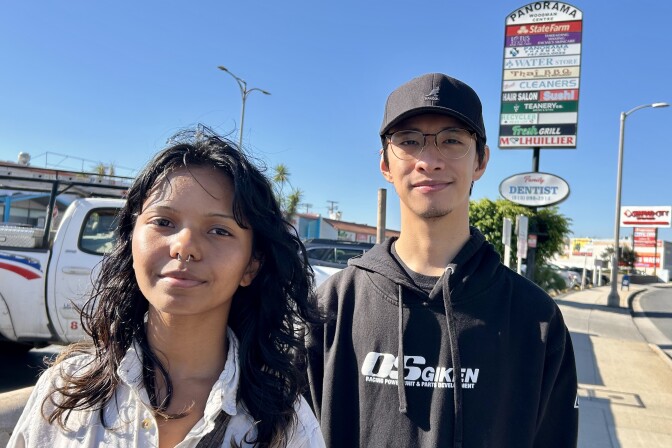 A male presenting person, wearing a black cap, and a female presenting person stand on a sidewalk.