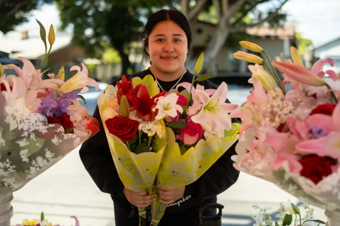 A young woman smiles as she holds two bouquets of flowers with more bouquets around her near a street.