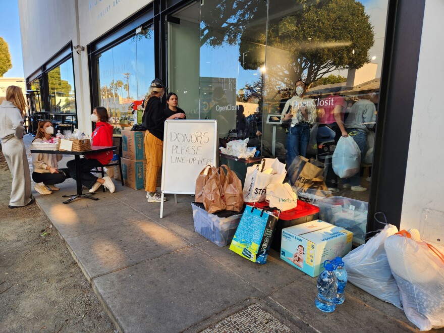 A street scene, in front of a series of glass store fronts. On the sidewalk is a pile of donated goods, like plastic bags of clothese and diaper boxes, next to a sign that says DONORS PLEASE LINE UP HERE
