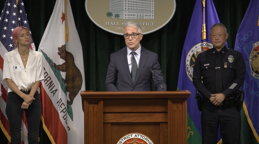 A screenshot of a press conference led by Los Angeles County District Attorney George Gascon (standing at the podium). He is flanked by a white woman with pink hair wearing a white shirt and black jeans, and an Asian man in an LAPD uniform.