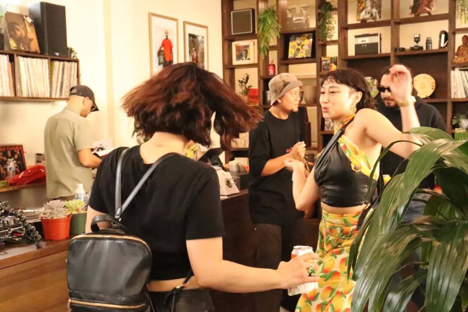 Two women in a shop are dancing. Behind them, there are two men chatting while a third man browses through records.