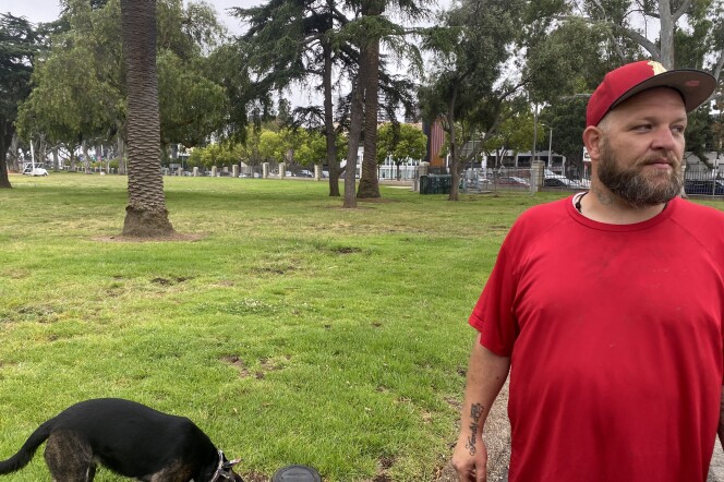 A tall bearded white man in a red baseball cap and red T-shirt stands on a lawn in front a grove of trees at the West Los Angeles VA campus. A small dog sniffs the grass behind him.