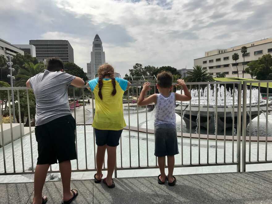 Three children stand at a railing looking over a fountain and L.A. City Hall with their backs to the camera.