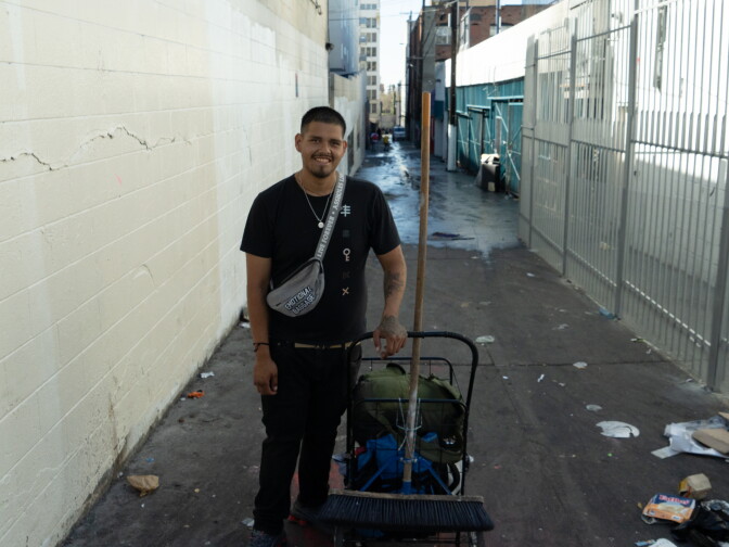 A smiling man in a black T-shirt and black pants stands in a narrow alley, holding a push broom beside a cart loaded with bags.