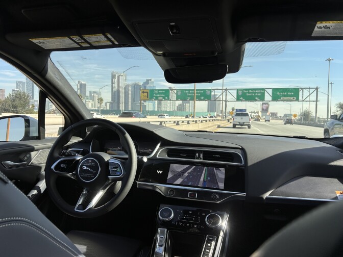 The interior of a car taken from the backseat, with notably no driver sitting behind the wheel. The car is driving on a freeway near the downtown Los Angeles skyline in the distance on the left.