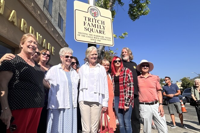 A crowd of people of varying ages are smiling and posing in front of a city street sign that reads "Tritch Family Square" and "Meeting the hardware needs of the Eagle Rock community since 1945" on a cloudless, sunny day.