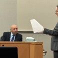 The setting is a courtroom: A man wearing a dark suit is sitting and looking at a man, also wearing a dark suit, as the man is speaking in reference to some papers in his hand. 