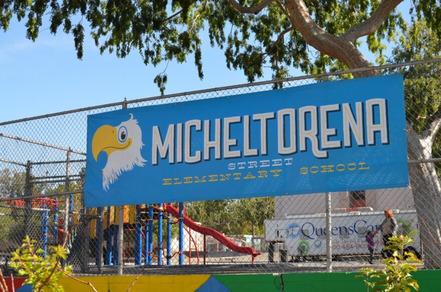 A bright blue sign that says "Michelotorena Street Elementary School," and has an illustrated eagle on it, hangs on a chain link fence outside a school playground.