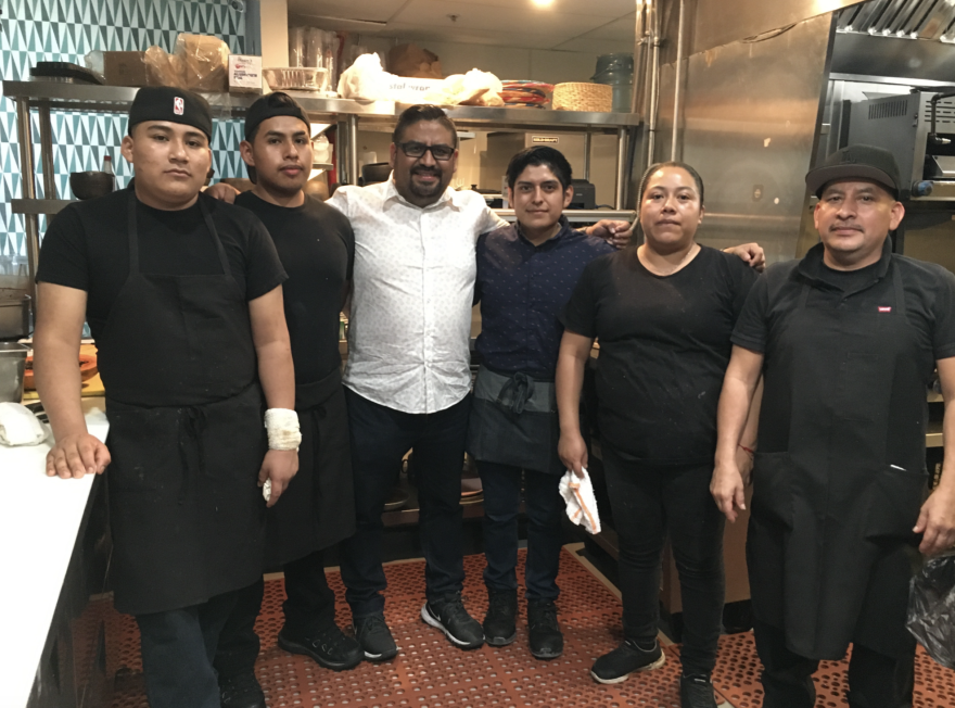 A group of kitchen staffers, all with varying brown skin tones, all wearing black, except for the restaurant owner, standing in the center, in a white shirt, inside a commercial kitchen.