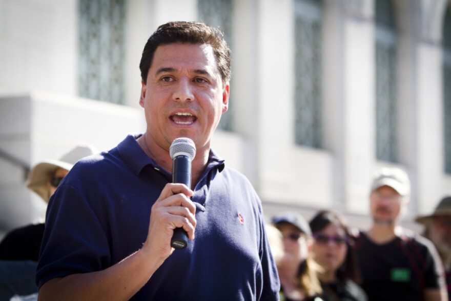 A man with medium tone skin wears a blue polo and holds a mic.