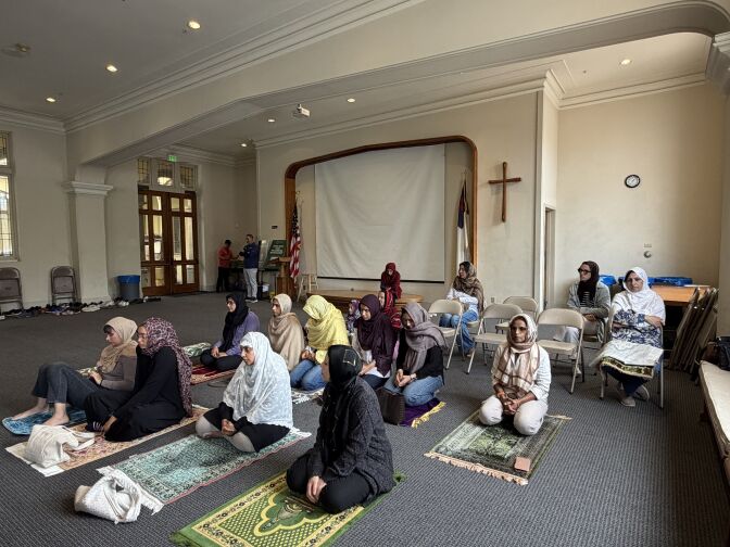 Women wearing headscarves sit on ornate prayer rugs. A cross hangs on the wall behind them.