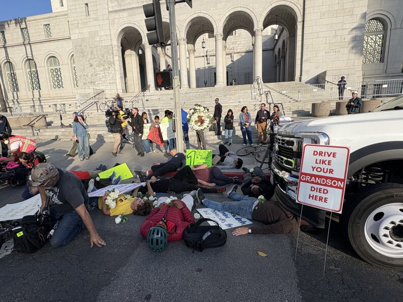 Several people lie in the street. There's a neon green sign sitting on the curb in the background that says "1 death x day." A sign in the foreground next to a truck reads "DRIVE LIKE YOUR GRANDSON DIED HERE."